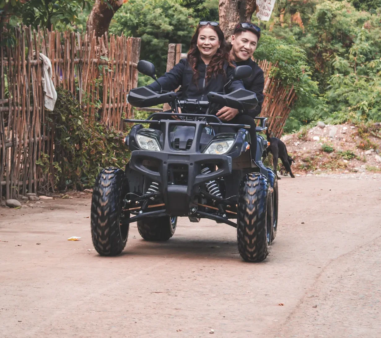 A couple rides an ATV on a dirt road bordered by green trees.