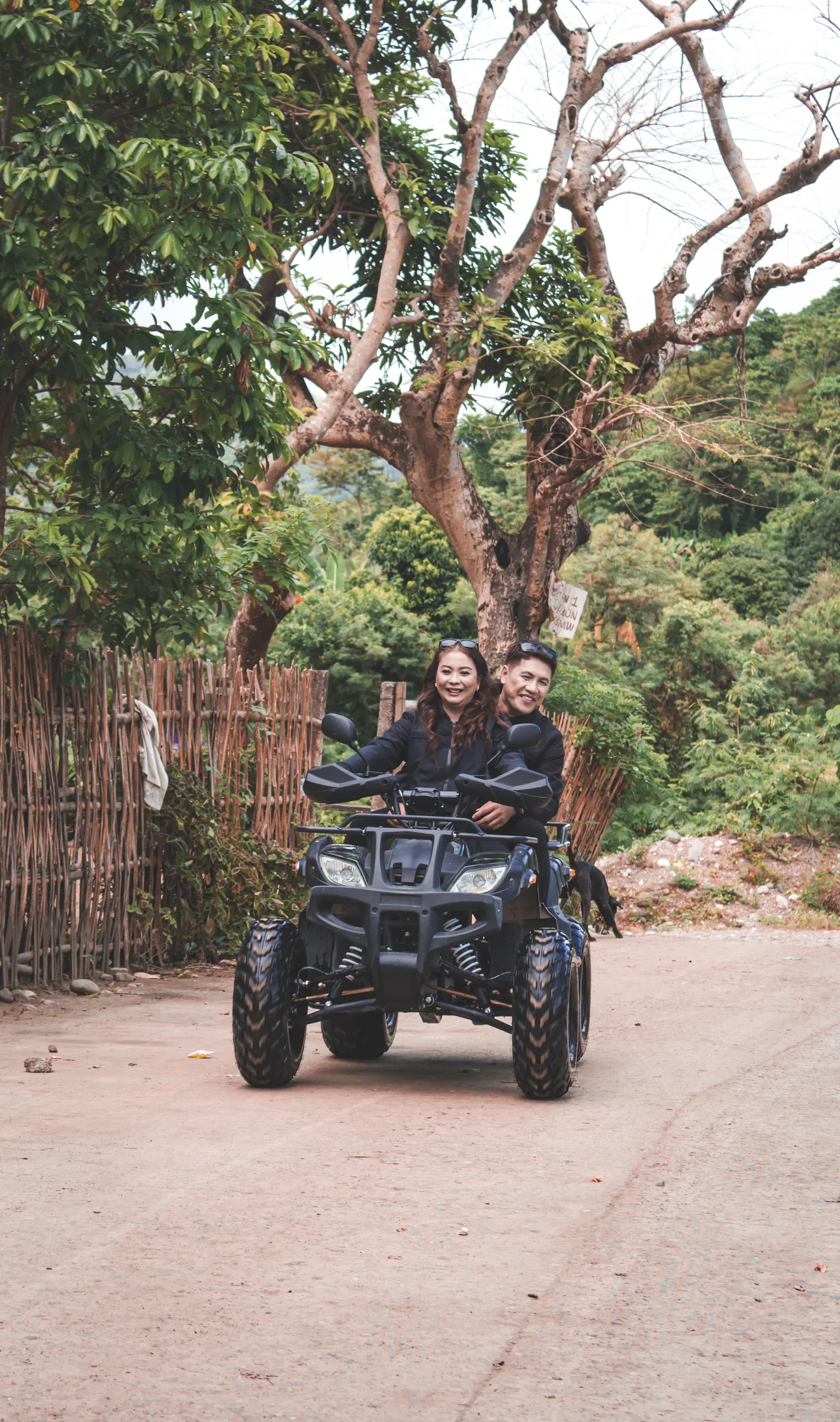 A couple rides an ATV on a tree-lined dirt road.